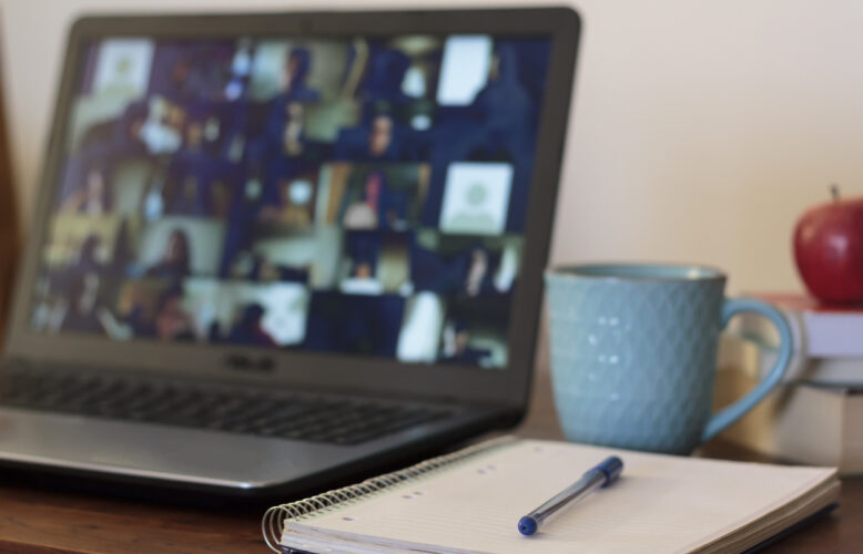 Open laptop shows blurred images of people attending virtual meeting. A notepad, pen, and coffee cup appear to the right.