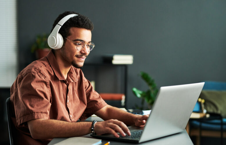 Young man with headphones looks at an open laptop at a desk.