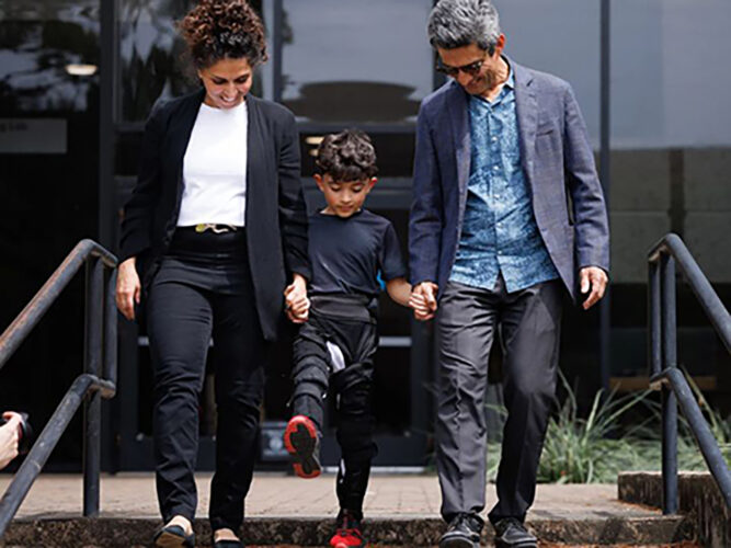 A woman and a man hold hands with a child between them who is wearing the MyoStep exoskeleton. All three are walking toward us.