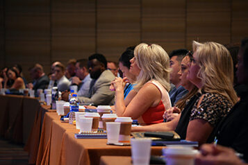 Professionals sit at a long table as they listen to a conference speaker.