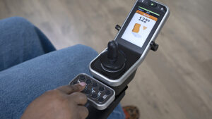 LiNX power chair electronics screen is visible with joystick. A person's hand is touching the programming controls.